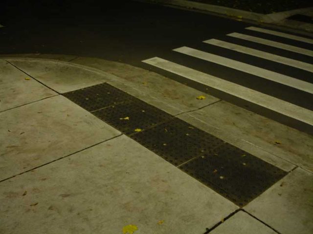 Donkere tegels met noppen op het trottoir naast een zebrapad, waar blinde mensen op kunnen voelen waar ze moeten oversteken.