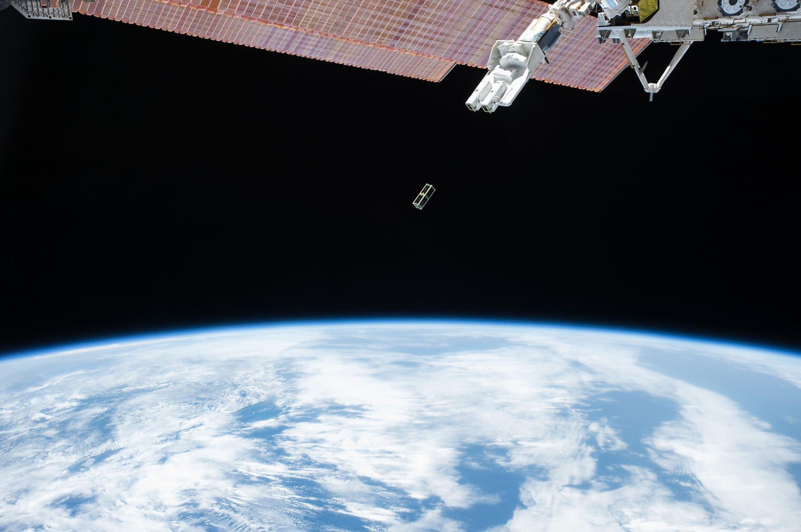 Een kleine satelliet zweeft in de ruimte boven de blauwe aarde met witte wolken, naast een deel van het ruimtestation.