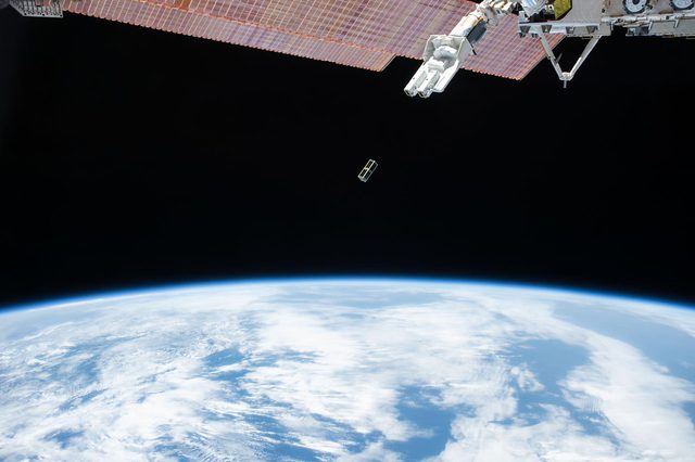 Een kleine satelliet zweeft in de ruimte boven de blauwe aarde met witte wolken, naast een deel van het ruimtestation.