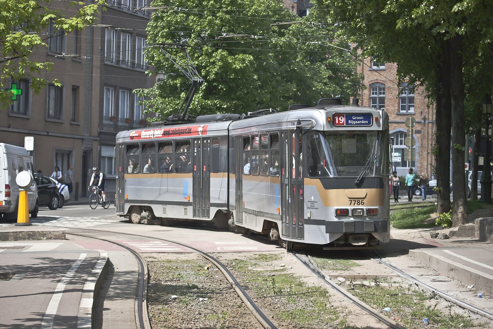 Een grijs met oranje tram van lijn 19 rijdt over de rails tussen bomen in een straat in Brussel.