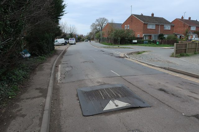 Een zwarte verkeersdrempel ligt op een rustige woonstraat met huizen en geparkeerde auto's op de achtergrond.