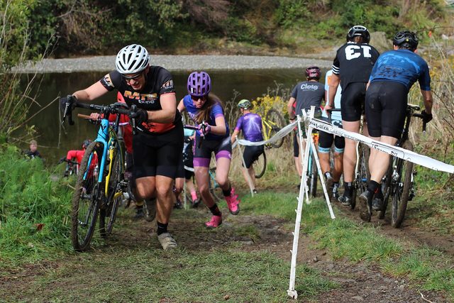 Veldrijders dragen hun fiets over een modderig pad langs het water tijdens een veldrijwedstrijd.