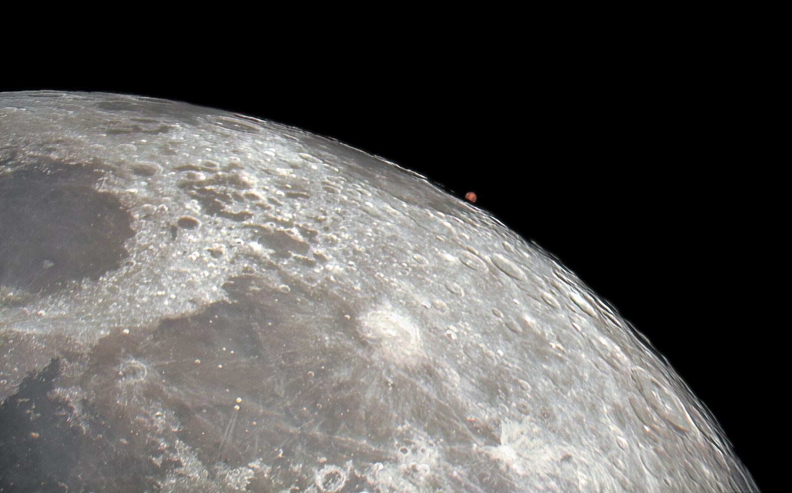 Een groot stuk van de kratermaan tegen een zwarte hemel, met rechts aan de rand een klein rood stipje: de planeet Mars.
