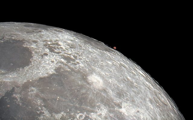 Een groot stuk van de kratermaan tegen een zwarte hemel, met rechts aan de rand een klein rood stipje: de planeet Mars.