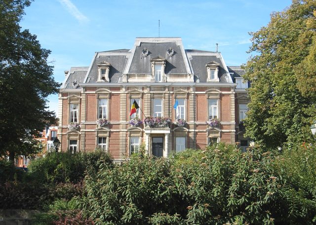Een groot oud bakstenen gemeentehuis met vlaggen en bloemen voor de ingang, omringd door groen.