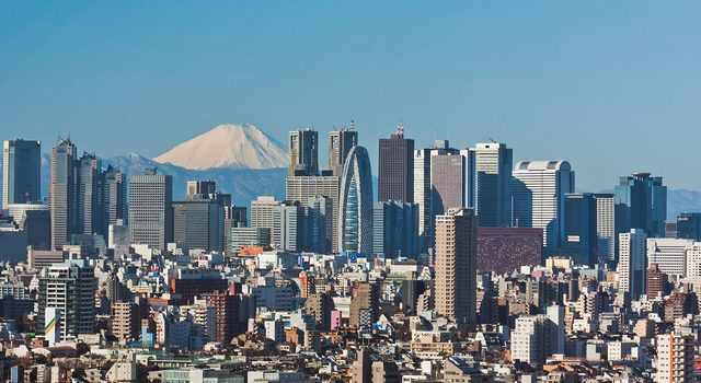 Hoge wolkenkrabbers en huizen van Tokio onder een blauwe lucht, met de besneeuwde berg Fuji ver achter de stad.