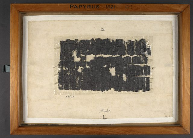 Een donker, verkoold stuk oude papyrus achter glas in een houten lijst.