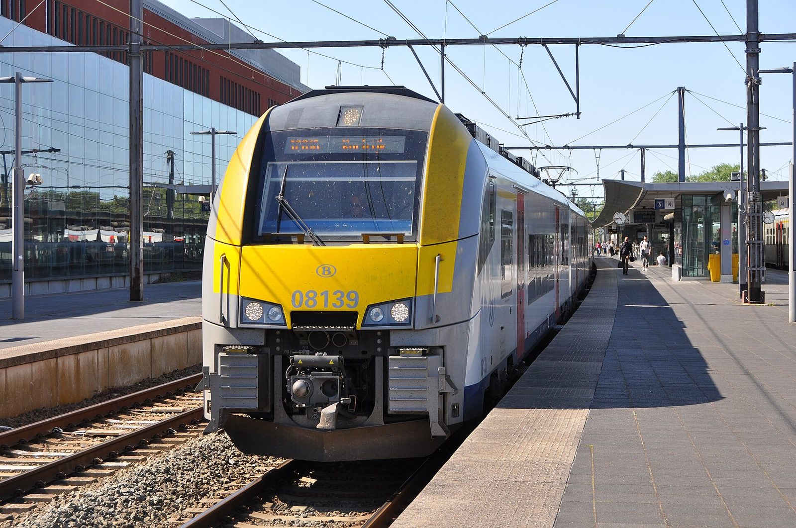 Een geel-witte NMBS-trein staat langs het perron in een station op een zonnige dag.