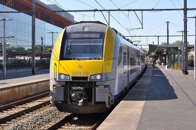 Een geel-witte NMBS-trein staat langs het perron in een station op een zonnige dag.