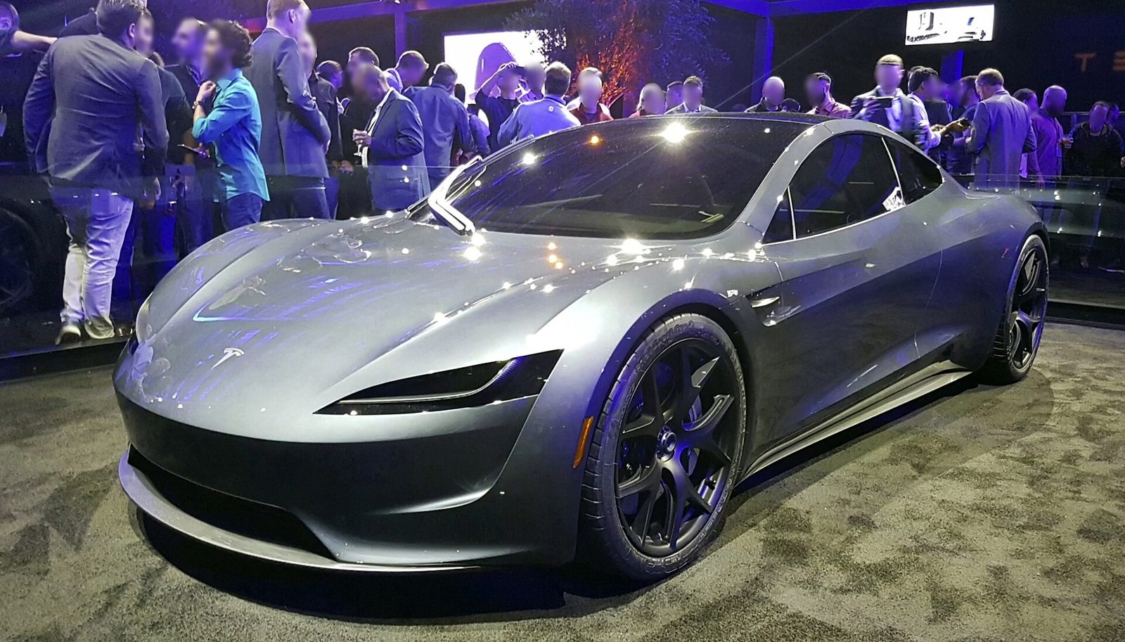 Een zilvergrijze Tesla Roadster staat op een podium met mensen op de achtergrond.