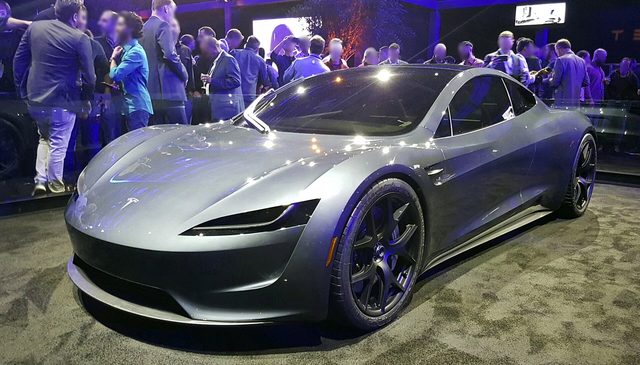 Een zilvergrijze Tesla Roadster staat op een podium met mensen op de achtergrond.