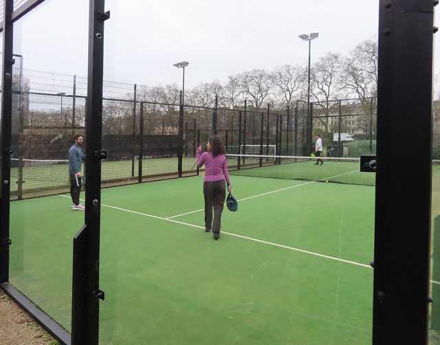 Een man en een vrouw staan met padelrackets op een groen padelterrein achter glazen wanden.