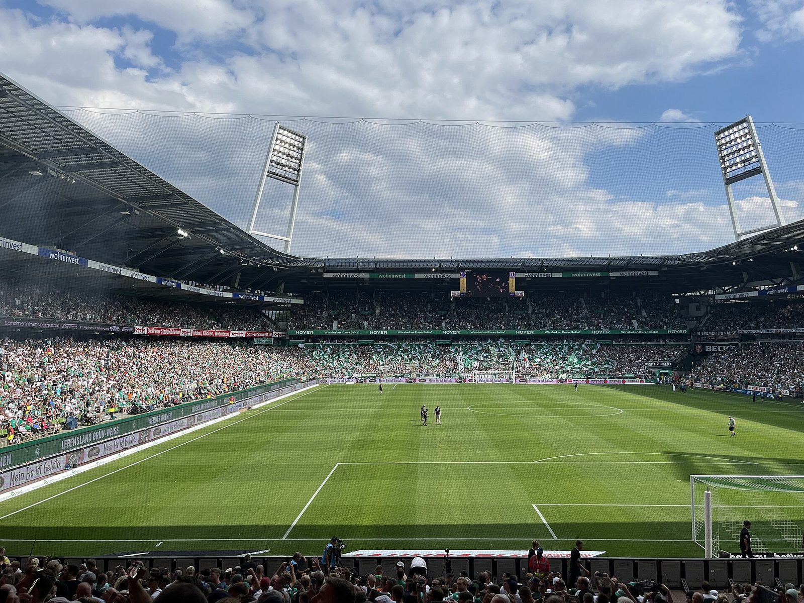 Een groen voetbalveld in een groot stadion met veel supporters op de tribunes onder een blauwe lucht.