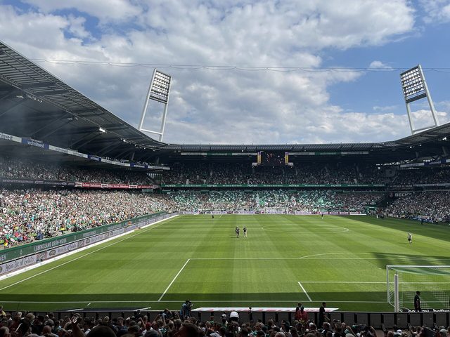 Een groen voetbalveld in een groot stadion met veel supporters op de tribunes onder een blauwe lucht.