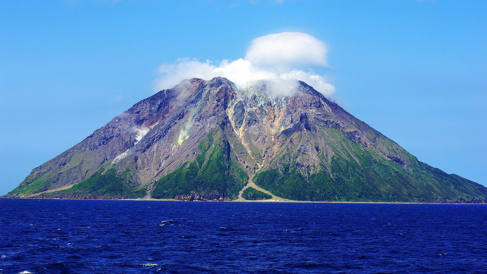 Een kale vulkaanberg met een wolkje bij de top, die uit de diepblauwe zee omhoog steekt.