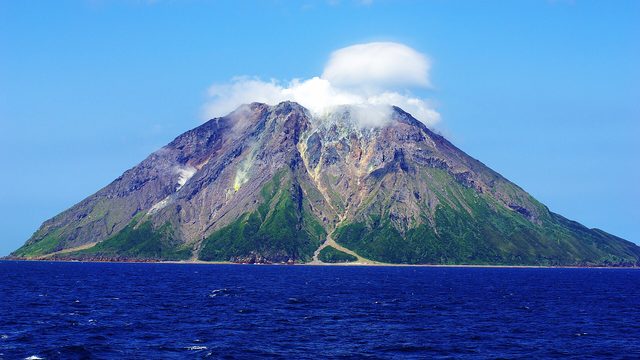 Een kale vulkaanberg met een wolkje bij de top, die uit de diepblauwe zee omhoog steekt.