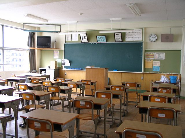 Een leeg klaslokaal met houten tafels en stoelen en een groen schoolbord vooraan.