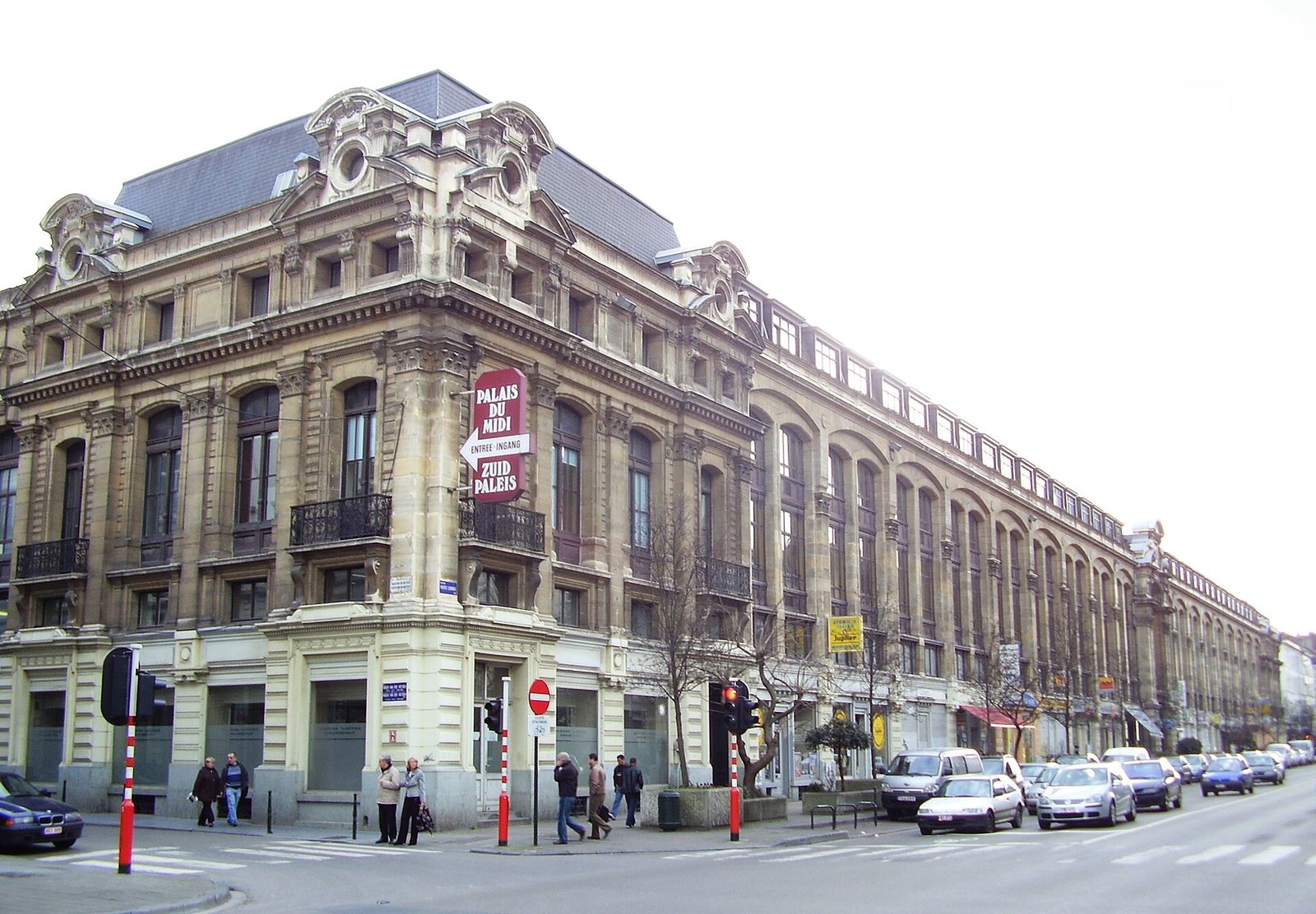 Een groot oud stenen gebouw op een straathoek in Brussel, met auto's en een paar voetgangers ervoor.