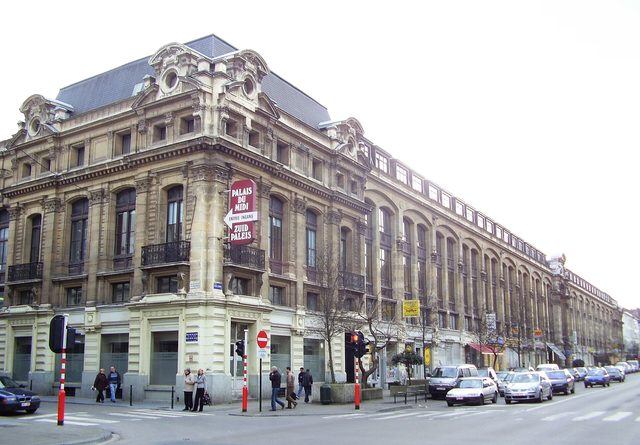 Een groot oud stenen gebouw op een straathoek in Brussel, met auto's en een paar voetgangers ervoor.