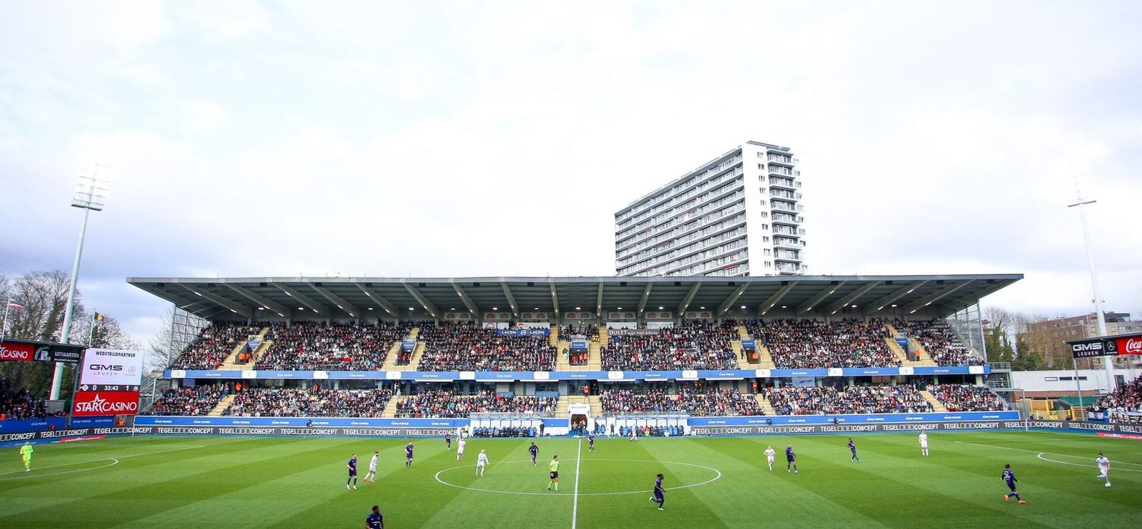 Een volle tribune van stadion Den Dreef met spelers op het grasveld tijdens een voetbalwedstrijd.