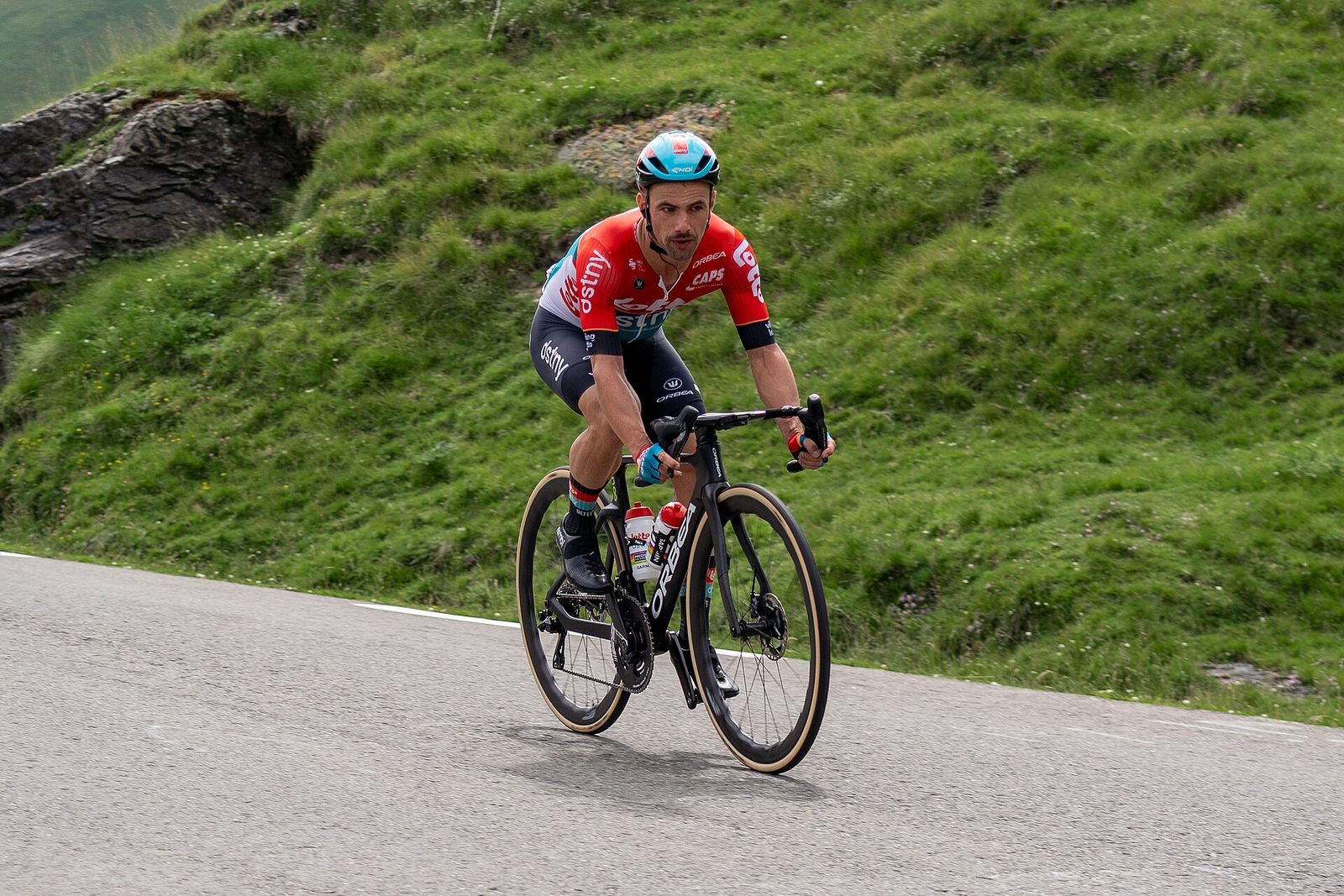 Een wielrenner in rood shirt en blauwe helm fietst snel een bergweg af, naast een grazige helling.