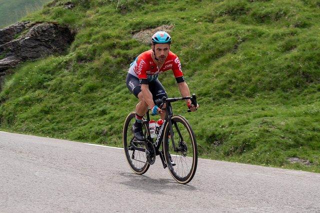 Een wielrenner in rood shirt en blauwe helm fietst snel een bergweg af, naast een grazige helling.