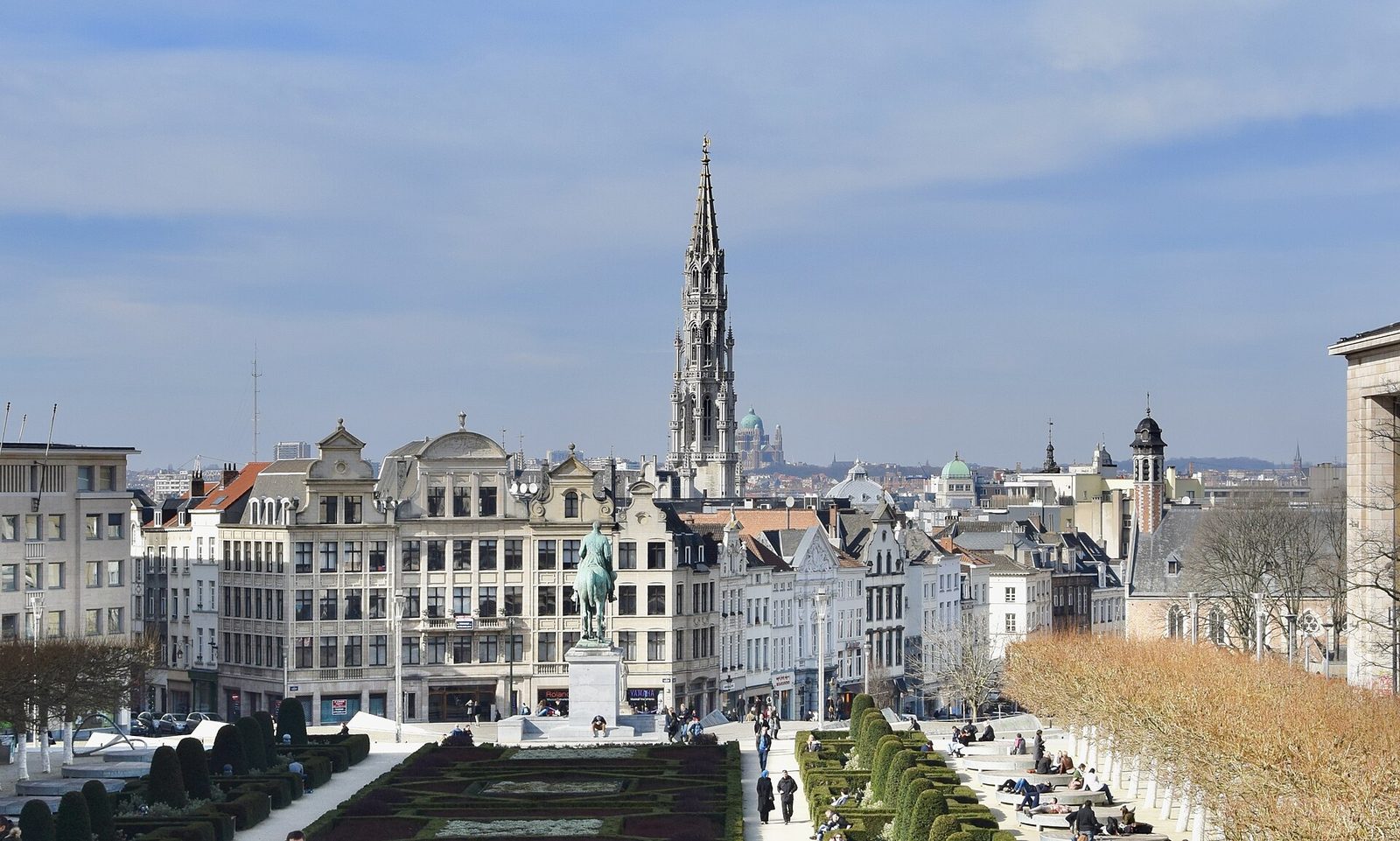 Uitzicht over Brussel met oude gebouwen, een hoge kerktoren en een tuin op de voorgrond onder een blauwe lucht.